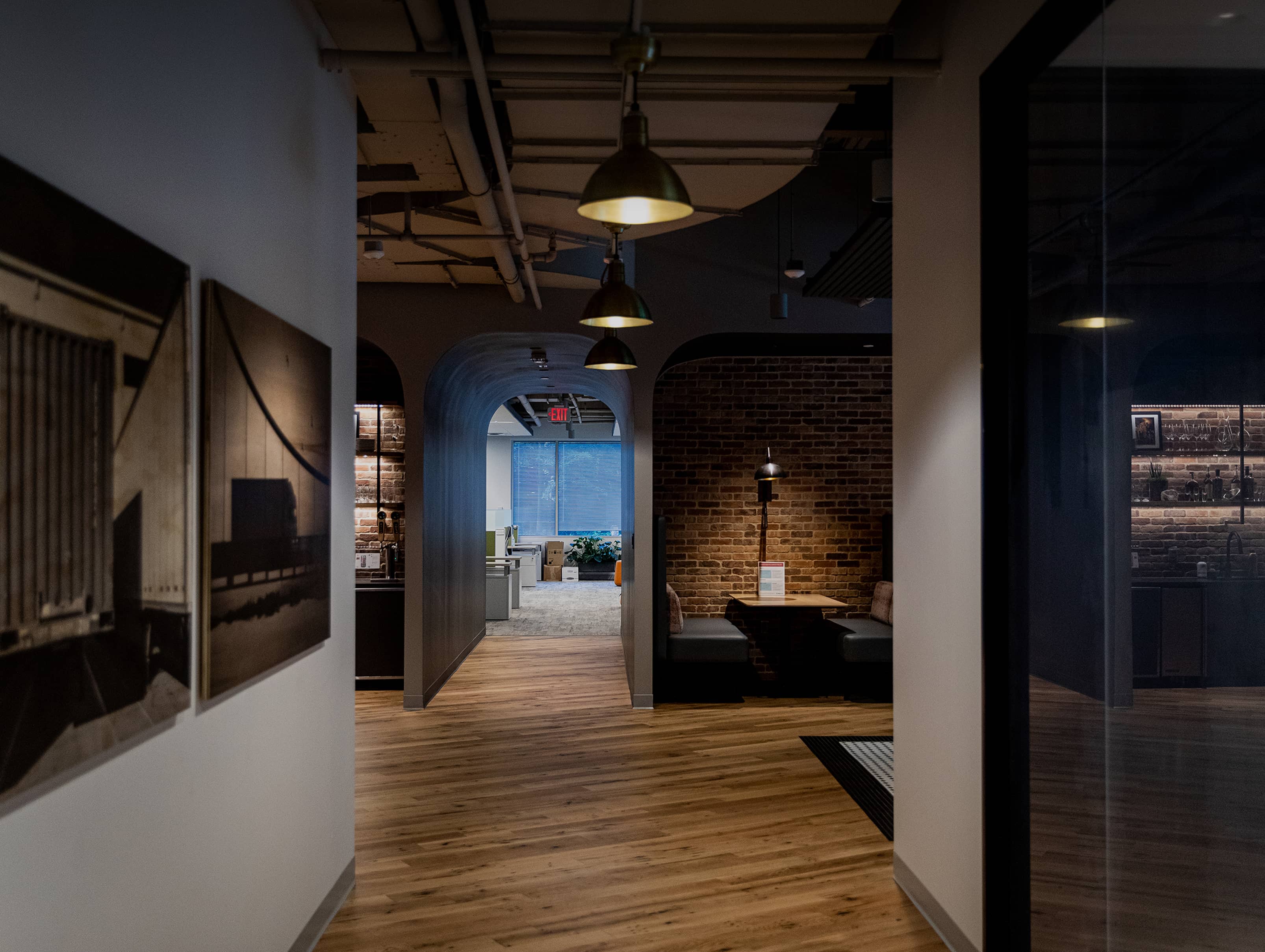 Dimly lit hallway with wooden floors and brick walls. Industrial-style lights hang from the ceiling. Artwork decorates the left wall.