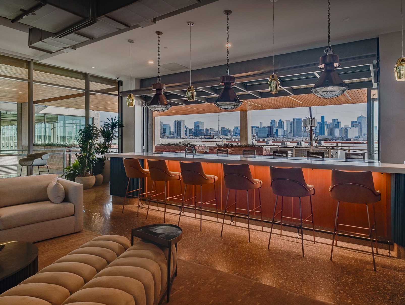 Sophisticated rooftop bar with a cityscape view, featuring modern pendant lighting, sleek counter with leather stools, and indoor plants. Urban elegance.