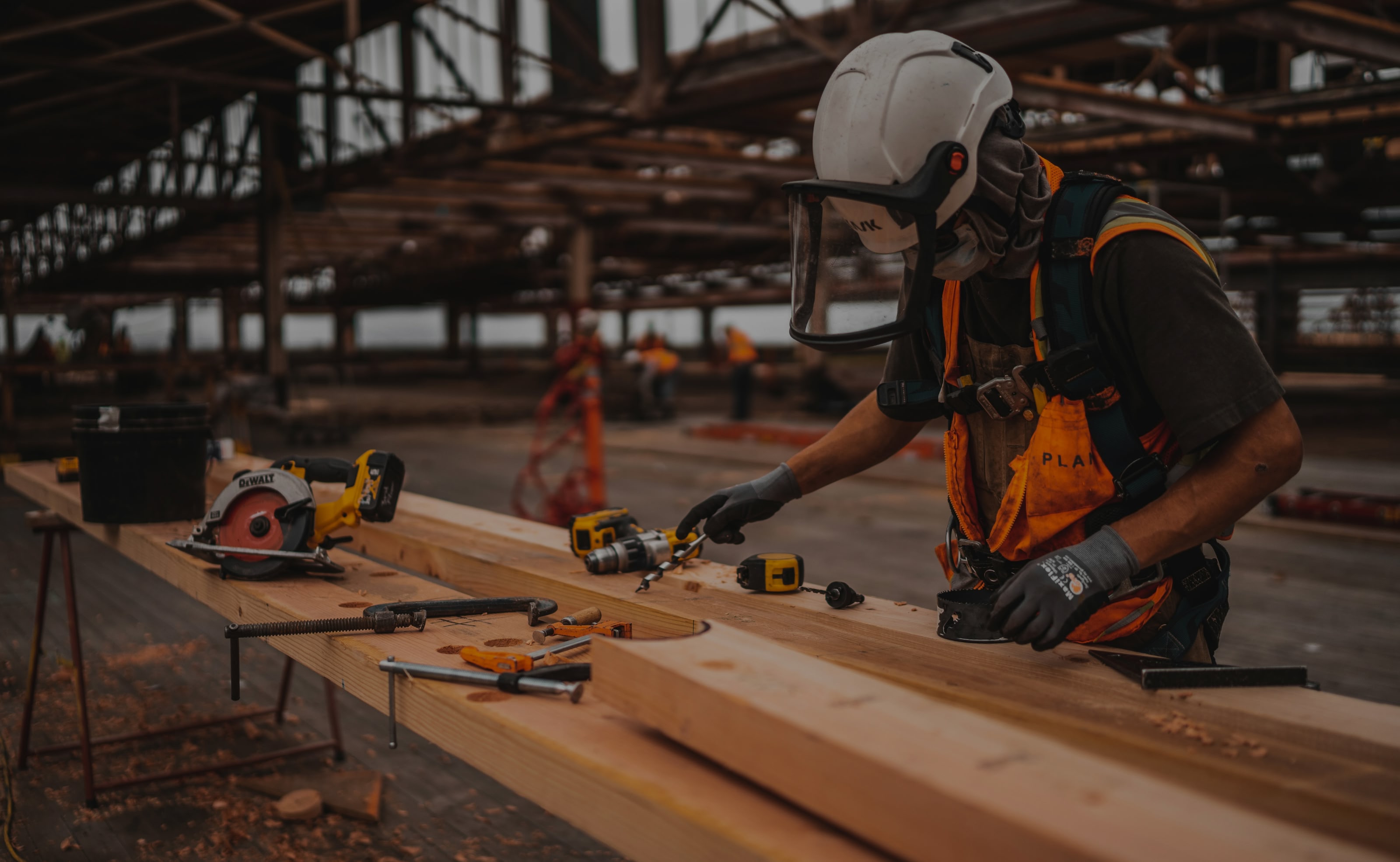 A construction worker in safety gear measures wooden beams on a table with various tools in an industrial site, conveying focus and craftsmanship.