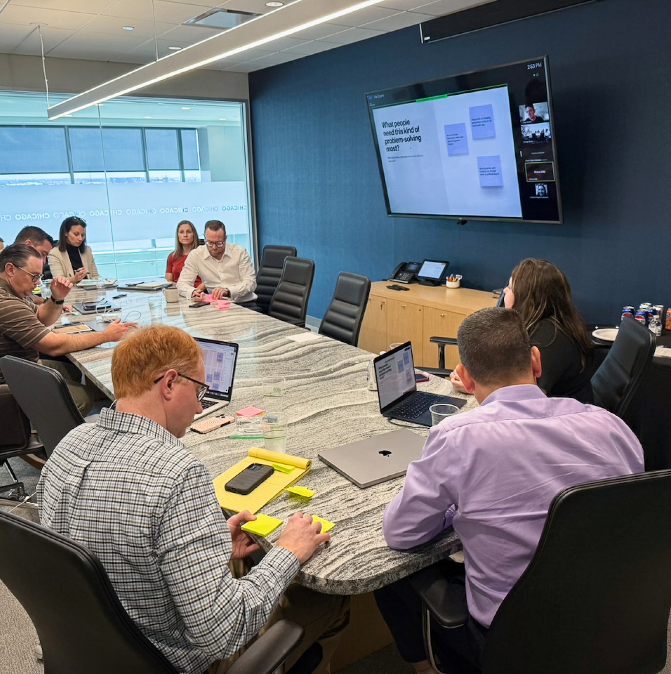 A group of eight people sit around a conference table with laptops and papers. A screen displays a presentation. The atmosphere is focused and collaborative.